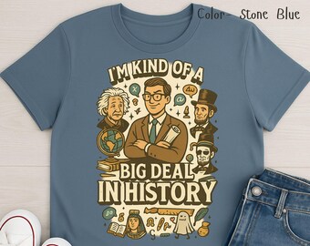 Può includere: T-shirt blu pietra con una grafica raffigurante un uomo dei cartoni animati con gli occhiali e la scritta "I'M KIND OF A BIG DEAL IN HISTORY". Il design include personaggi storici.