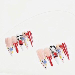 May include: A set of stiletto-shaped false nails with a clown theme. The nails feature a variety of designs, including red and white stripes, clown faces, and polka dots. The color palette includes red, white, pink, and yellow.