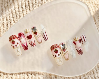 May include: A set of artificial nails with various designs, including strawberries, hearts, crowns, and geometric patterns. The nails feature a mix of red, white, gold, and pink colors, with some adorned with rhinestones and bows. The nails are arranged on a white, heart-shaped plate.