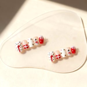 Peut inclure: Ensemble de faux ongles sur le thème de Noël. Chaque ongle présente des motifs festifs, notamment des cannes en sucre, des croix rouges et blanches, des bonhommes de neige et des accents rouges. Les ongles sont disposés sur une surface transparente de forme ovale.