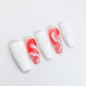 Op de afbeelding: Vijf valse nagels in een doodskistvorm. Twee nagels hebben een rood-wit wervelontwerp, terwijl de andere drie een wit, getextureerd patroon hebben. De nagels zijn geplaatst tegen een effen witte achtergrond.