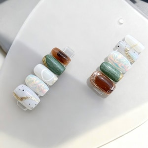 Può includere: Una collezione di unghie finte con vari design. Le unghie presentano colori come bianco, oro, verde, marrone e finiture iridescenti. Alcune unghie hanno accenti glitterati, mentre altre presentano motivi e texture.