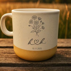 Puede incluir: Taza de cerámica color crema con base amarilla, con un diseño floral y las iniciales "h♡k" con la fecha "10.11.2025". La taza tiene una textura moteada y un asa curva, sobre un fondo borroso al aire libre.