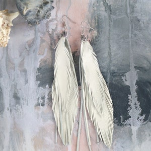 以下が含まれることがあります： 銀色のフックが付いた、長い白い革の羽根のイヤリングのペア。イヤリングは、質感のある灰色の背景にぶら下がっています。