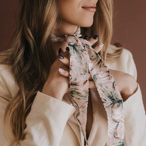 May include: A long, narrow neck scarf featuring a light pink background with a dark green and black tropical leaf pattern. The scarf is tied loosely around the neck, with two long, flowing ends hanging down over a cream-coloured double-breasted blazer.