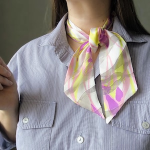 Op de afbeelding: Een kleurrijke zijden sjaal met een levendig abstract ontwerp in roze, gele en witte tinten is om de nek geknoopt. De sjaal wordt gecombineerd met een blauw-wit gestreept overhemd, wat een stijlvolle uitstraling creëert.
