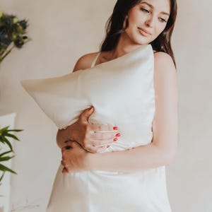 May include: A cream-colored silk pillow is held in front of a person. The pillow has a smooth, soft texture. The person is wearing a matching cream-colored silk nightgown. The background is a neutral, light color.