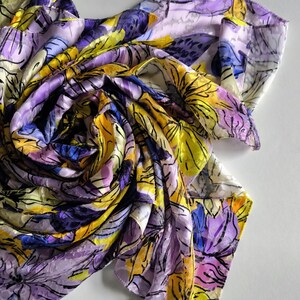 Op de afbeelding: Een sjaal met bloemenpatroon in paars, geel en blauw. Het ontwerp heeft een aquarel-stijl met zwarte contouren, wat een levendig en artistiek accessoire creëert. De stof lijkt licht en vloeiend.