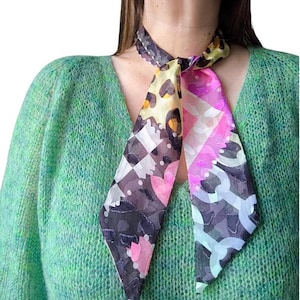 Op de afbeelding: Een kleurrijke zijden sjaal met verschillende patronen, waaronder luipaardprint, geometrische vormen en bloemmotieven. De sjaal heeft een mix van kleuren, waaronder roze, paars, geel en zwart. De sjaal is om de nek geknoopt.