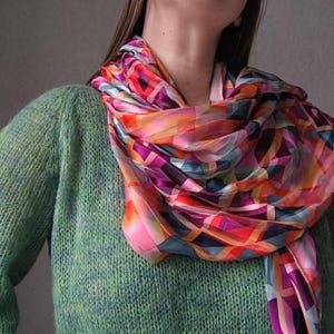 Puede incluir: Un pañuelo de seda colorido con un diseño geométrico en tonos rosa, naranja, morado y azul. El pañuelo está drapeado sobre un suéter verde, creando un contraste vibrante. El diseño del pañuelo presenta una mezcla de formas y líneas.