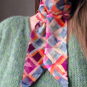 Op de afbeelding: Een kleurrijke zijden sjaal met een geometrisch patroon in tinten oranje, roze, paars en blauw. De sjaal is om de nek geknoopt, wat een stijlvol accessoire creëert. Het ontwerp van de sjaal bestaat uit elkaar kruisende lijnen en vormen.