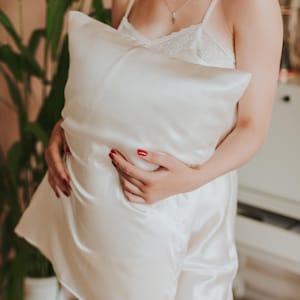 May include: A white satin pillow is held in front of a person wearing a matching satin pajama set. The pillow has a smooth, lustrous surface. The pajama set includes a camisole and shorts with lace trim. Red nail polish is visible.