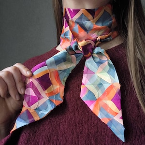 Op de afbeelding: Een kleurrijke zijden sjaal met een geometrisch patroon in tinten blauw, oranje, paars en roze. De sjaal is om de nek geknoopt, met de uiteinden naar beneden. Een levendig accessoire.
