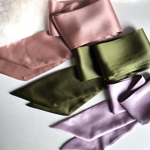 May include: A selection of silk scarves in dusty rose, olive green, and lavender. The scarves are displayed on a white surface, with a soft, white, textured material in the background. The scarves have a smooth, shiny texture.