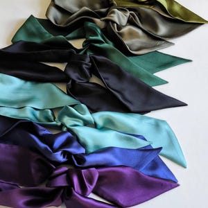 Può includere: Una collezione di sei nastri per capelli in raso disposti in fila verticale, caratterizzati da una sfumatura di colori che include verde oliva, verde bosco scuro, nero, verde acqua, blu reale e viola intenso, ciascuno annodato con punte.