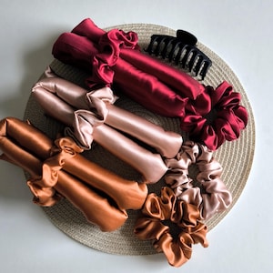 Può includere: Tre arricciacapelli in raso nei colori rosso scuro, rosa chiaro e rame, ciascuno abbinato a un elastico in raso coordinato. È inclusa una pinza per capelli in plastica nera, caratterizzata da superfici in tessuto liscio e lucido con estremità arricciate.
