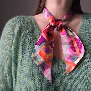Op de afbeelding: Een kleurrijke zijden sjaal met een geometrisch patroon in roze, paars, oranje en blauw is om de nek geknoopt. De sjaal wordt gecombineerd met een lichtgroene, getextureerde trui. Het ontwerp van de sjaal bevat gebogen en hoekige vormen.