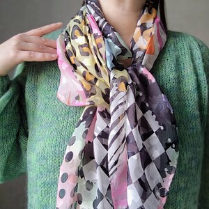 Könnte beinhalten: Ein farbenfrohes Seidentuch mit verschiedenen Mustern, darunter Leopardenmuster, Tupfen und ein Schachbrettmuster. Der Schal hat eine Mischung aus Farben, darunter Rosa, Gelb, Schwarz und Weiß. Der Schal ist um den Hals drapiert.