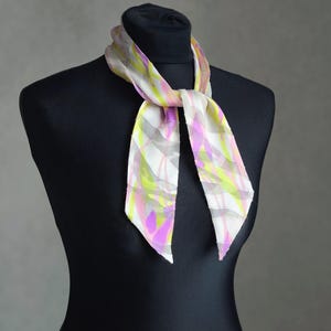 Op de afbeelding: Een kleurrijke zijden sjaal met een abstract patroon. De sjaal heeft een mix van wit, roze, geel en grijs. De sjaal is om een zwarte mannequin geknoopt, wat het ontwerp en de drapering benadrukt.