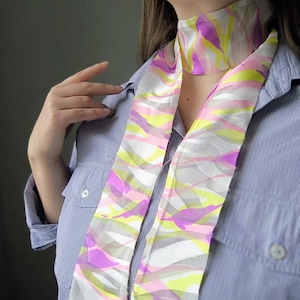 May include: A colorful silk scarf with a vibrant abstract pattern in shades of pink, yellow, and purple on a white background. The scarf is long and narrow, designed to be worn around the neck. The scarf is paired with a light blue button-down shirt.