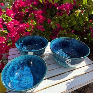 May include: Three blue ceramic bowls with a glossy, textured finish. The bowls are a deep shade of blue with lighter blue and white accents. They are arranged on a white slatted table, with vibrant pink flowers in the background.