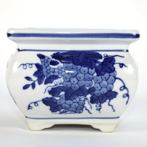 May include: A white ceramic planter with a blue grape design. The planter has a square top and rounded sides, with a blue stripe around the rim. The grape design features clusters of grapes and leaves.