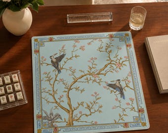 May include: A square light blue mat featuring a central illustration of a brown tree with pink blossoms and three birds. The mat is framed by a decorative border with geometric patterns and red floral accents in each corner of the design.
