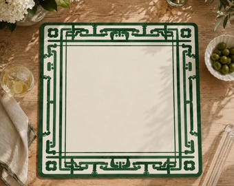 May include: A square placemat featuring a solid cream-colored center framed by a dark green border with a geometric, interlocking pattern. The design consists of straight lines and stylized corner motifs that create a structured, symmetrical frame.