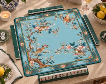 May include: A square mahjong mat featuring a light blue surface with a detailed illustration of birds perched on flowering branches. The mat is bordered by a decorative floral pattern and is surrounded by four dark green tile racks and tiles.