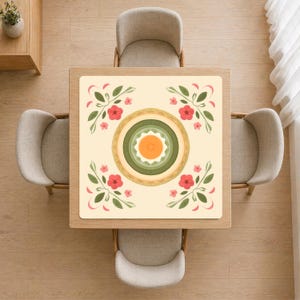 Può includere: Vista dall'alto di un tavolo da pranzo quadrato con un sottopiatto floreale. Il sottopiatto ha uno sfondo color crema con un motivo circolare centrale nei toni del verde, arancione e oro, circondato da fiori rossi e foglie verdi.