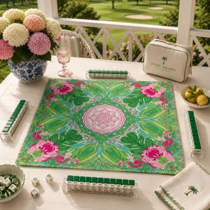 May include: A square mahjong mat featuring a vibrant green and pink floral pattern with a central mandala design. The set includes four clear tile racks, white mahjong tiles with green backs, two dice, a white storage bag labeled HOME AMARIS, and a napkin.