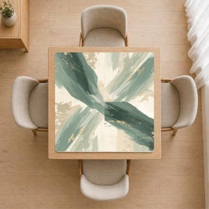 Può includere: Vista dall'alto di un tavolo quadrato in legno con un sottopiatto decorativo con un design astratto nei toni del verde, beige e oro. Quattro sedie beige circondano il tavolo, posto su un pavimento in legno chiaro. È visibile una piccola pianta.