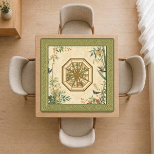 May include: Overhead view of a square wooden table with a decorative floral and bird-themed table mat. The mat features a green border and a central octagonal design. Four beige chairs surround the table, set on a light wood floor.