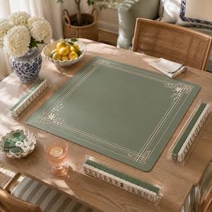 May include: A square sage green mahjong mat featuring a thin gold border with decorative corner floral motifs. The set includes three clear acrylic tile racks filled with green and white mahjong tiles, arranged on a wooden table surface.