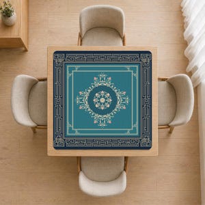 Peut inclure: Vue aérienne d'un tapis de table carré turquoise avec un motif floral et géométrique orné. Le tapis est entouré d'une bordure bleu foncé et est centré sur une table en bois clair. Quatre chaises beiges entourent la table.