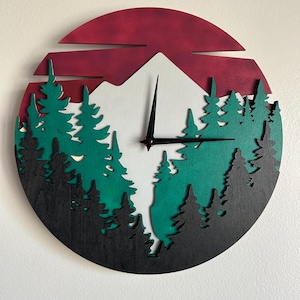 Peut inclure: Une horloge murale ronde en bois représentant une scène de montagne et de forêt. Le cadran de l'horloge présente un soleil rouge, une montagne blanche et des arbres verts et noirs. Les aiguilles de l'horloge sont noires.