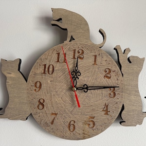 Puede incluir: Reloj de pared de madera con temática de gatos y acabado marrón claro. La esfera del reloj es redonda con un diseño texturizado y tres siluetas de gatos. Las manecillas del reloj son negras y el segundero es rojo.