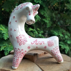 Può includere: Statuetta di cavallo in ceramica bianca con motivi decorativi rossi e il nome "Ann". Il cavallo ha un collo curvo e sta su quattro zampe. I motivi includono disegni floreali e geometrici.