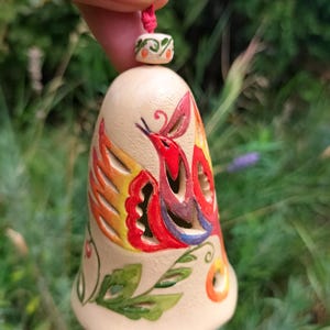 May include: A hand-painted ceramic bell with a bird design. The bell features a cream-colored base with a vibrant bird illustration in red, orange, yellow, and blue. A red cord and decorative bead are attached to the top. The bell is approximately 3 inches tall.