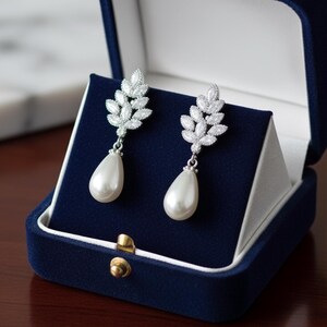 May include: A pair of silver-toned drop earrings with a leaf design and a pear-shaped pearl. The earrings are displayed in a dark blue velvet jewelry box with a gold clasp. The box is open, revealing the earrings against a dark blue background.