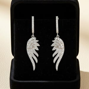May include: A pair of silver-toned dangle earrings in a black jewelry box. Each earring features a wing design, encrusted with small, sparkling stones. The earrings are displayed against a black velvet background, highlighting their intricate details and shine.