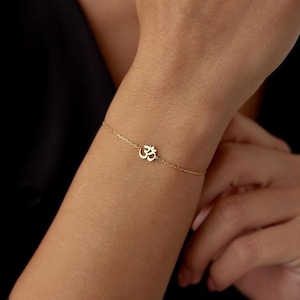 May include: A delicate gold-colored bracelet featuring the Om symbol. The bracelet is a fine chain with the Om symbol as a charm. The bracelet is worn on a wrist, and the background is a dark color.