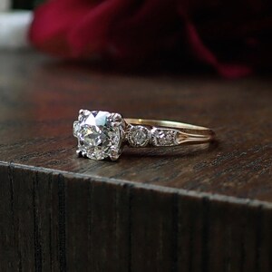 May include: A gold engagement ring with a large, round-cut diamond center stone. The ring features smaller diamonds on the band and is set against a dark wooden surface. The ring is a classic design.