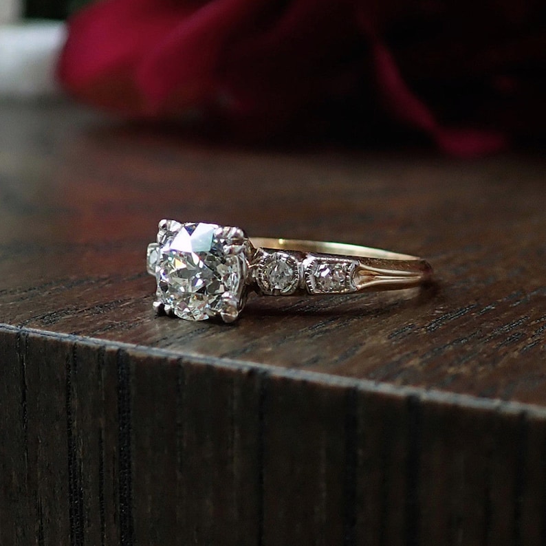 May include: A gold engagement ring with a large, round-cut diamond center stone. The ring features smaller diamonds on the band and is set against a dark wooden surface. The ring is a classic design.