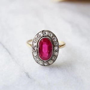 May include: A gold ring featuring an oval, vibrant red gemstone surrounded by a halo of smaller, clear stones. The ring's band is a simple gold design, and the setting is a mix of gold and silver tones.
