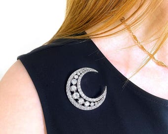 May include: A silver-toned crescent moon brooch, adorned with two rows of sparkling gemstones. The brooch is set against a black garment. A delicate gold chain is visible in the background.