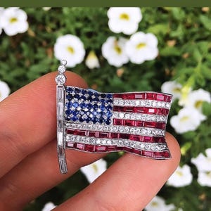 May include: A silver pin in the shape of an American flag, with red rubies and blue sapphires for the stripes and stars. The pin is set on a white background.