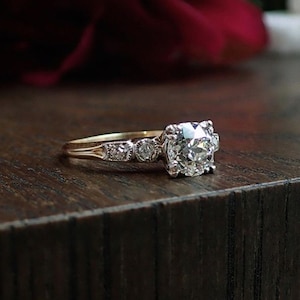 May include: A vintage-style engagement ring with a large, sparkling diamond center stone. The ring features a gold band with smaller accent diamonds. The ring is set on a dark wooden surface, with a blurred red background.