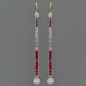 May include: A pair of elegant drop earrings. Each earring features a gold-colored hook, a top and bottom round clear stone, and a central column with alternating red and clear stones. The earrings are set against a gray background.