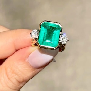 May include: A gold ring featuring a large, rectangular emerald gemstone with two smaller, round, clear gemstones on either side. The emerald is a vibrant green color, and the ring has a classic, elegant design.